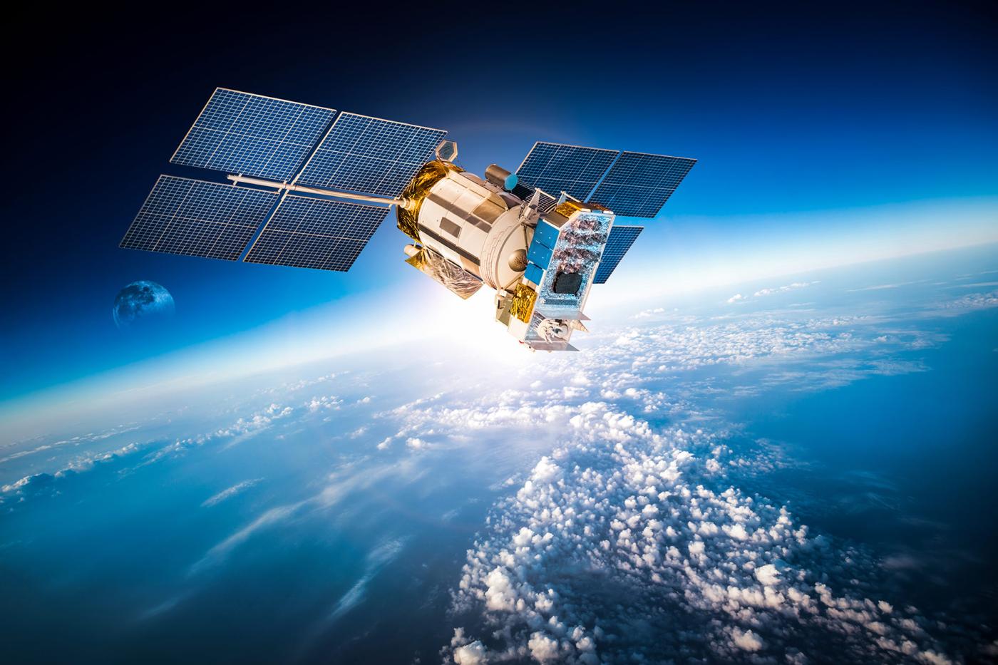 Satellite orbiting Earth with large solar panels extended, above a layer of white clouds and blue atmosphere, with the Moon visible in the distant background.